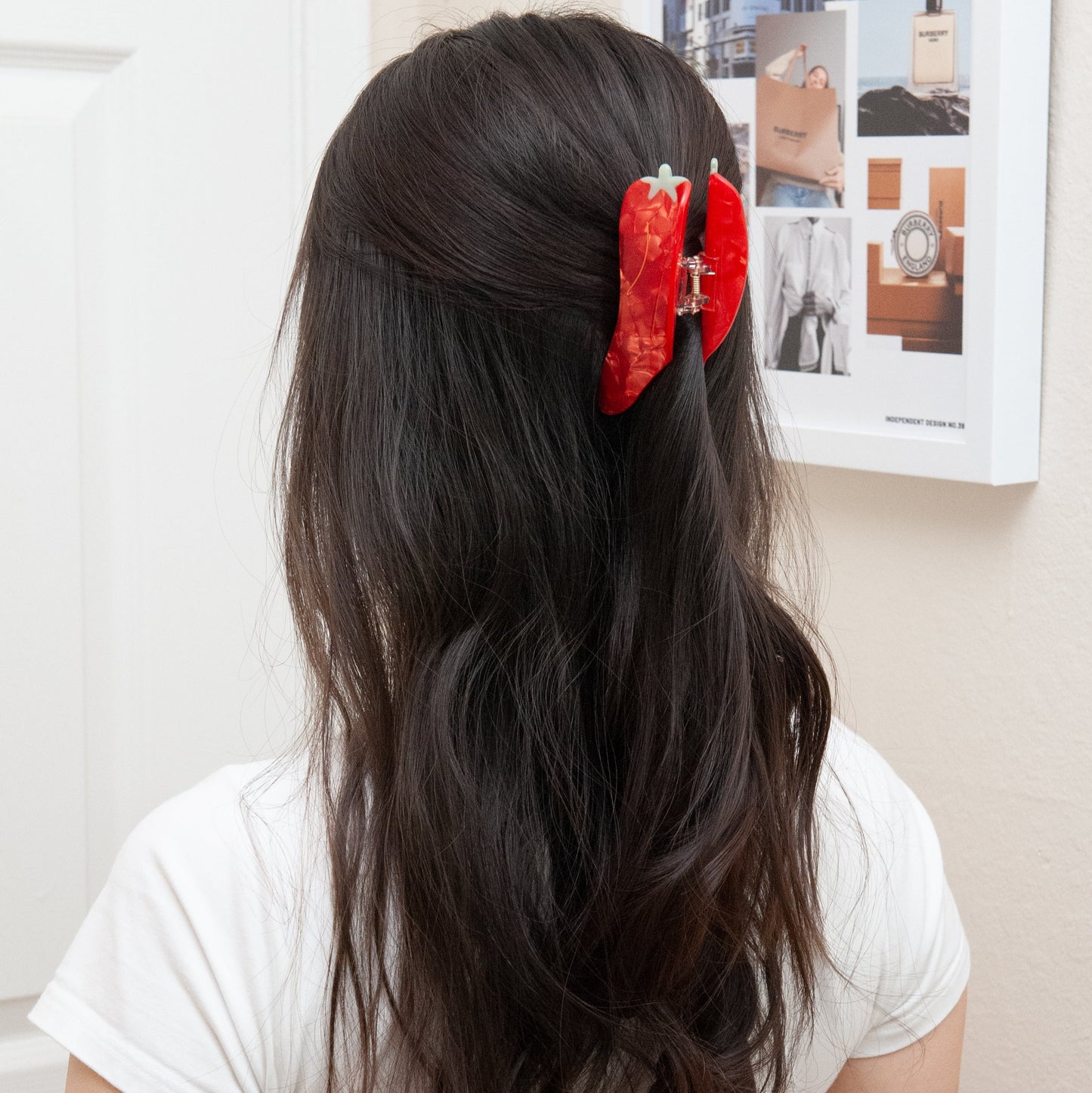 Person with long dark hair wearing a red chili hair clip, standing in front of a wall with a poster.