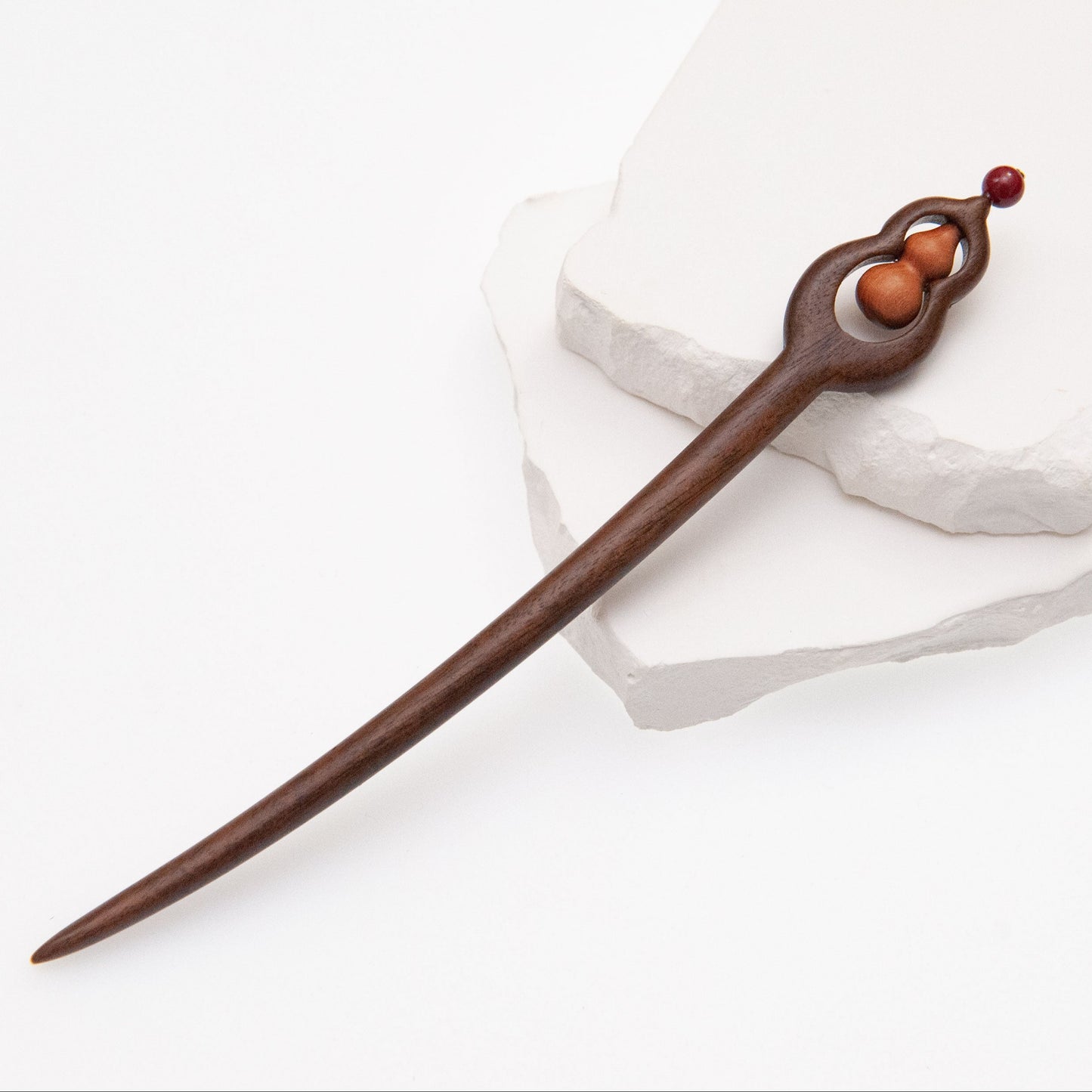 A wooden hair stick with a gourd bottle shape, featuring a red decorative element at the top, resting on a white stone.