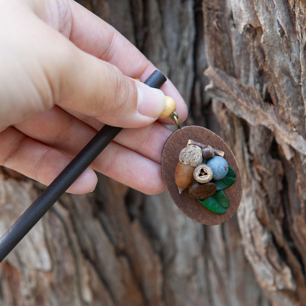Handcrafted wooden hair stick with small wooden beads, dried acorns, coffee beans, and blueberries, evoking a whimsical forest vibe.