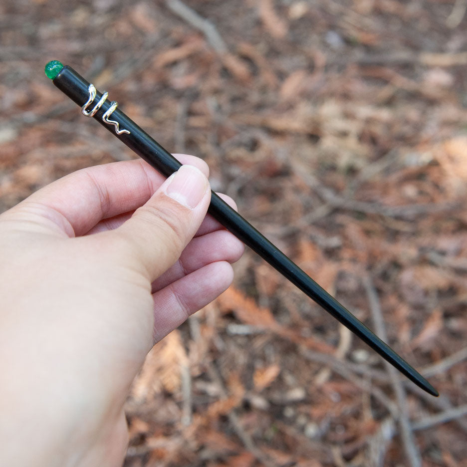 Handmade ebony hair stick with a silver snake coiled around it and a small green bead at the end, elegant and mysterious design.