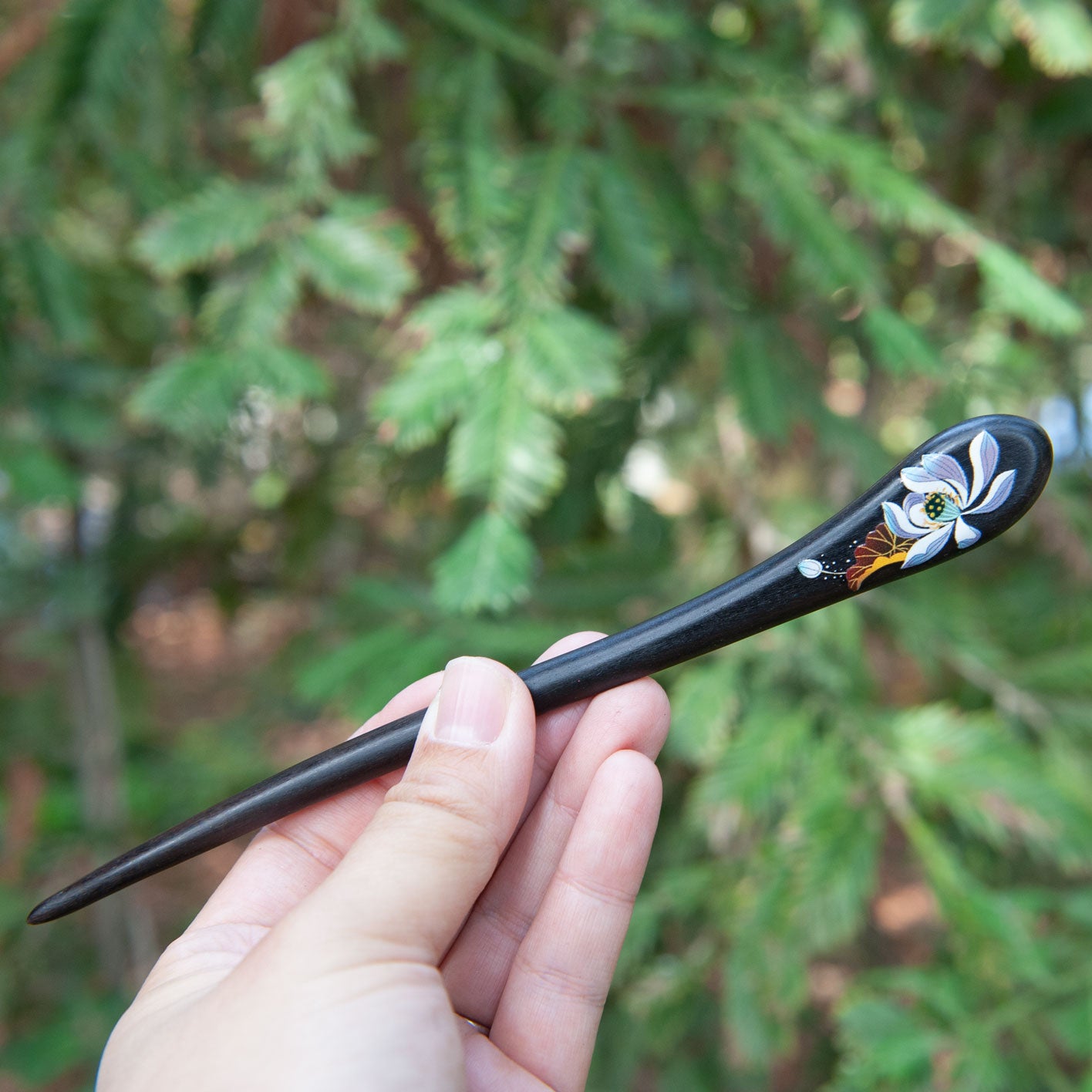 Hand holding a Chinese style black hairpin with floral design against a green foliage background