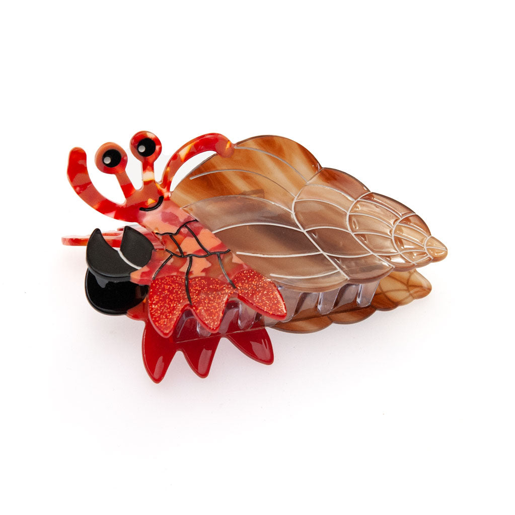 A decorative hair claw clip shaped like a hermit crab, featuring a translucent brown and amber shell, red glittery claws, and black accents, shown on a plain white background.