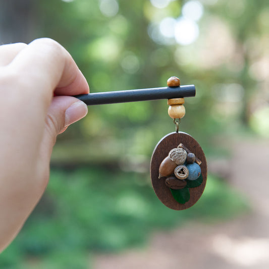 Hand holding a decorative hair stick with nature-themed beads against a blurred green background